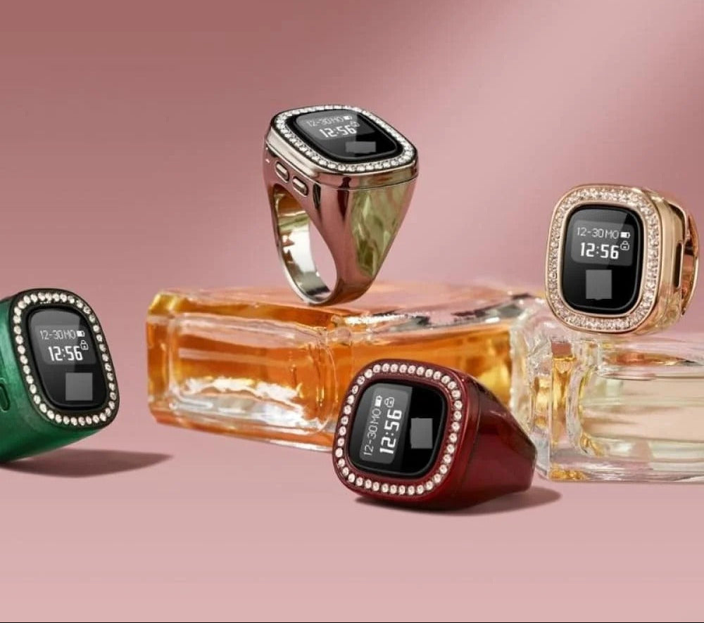 Four smartwatch rings displayed on and around perfume bottles against a soft pink studio background. The rings have rectangular black digital screens, crystal-like borders, and different band colors including rose gold, gold, dark red, and green.