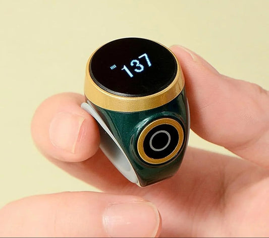 A close-up of a hand holding a small smart ring with a dark green body, gold trim, and a black circular display showing the number 137. The background is a solid pale yellow.