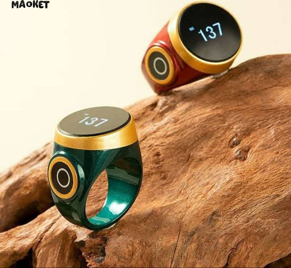 Two smart rings displayed on a piece of natural wood. One ring is dark green with gold trim and a black screen showing '137'; the other is red with gold trim and a black screen showing '10:37'.