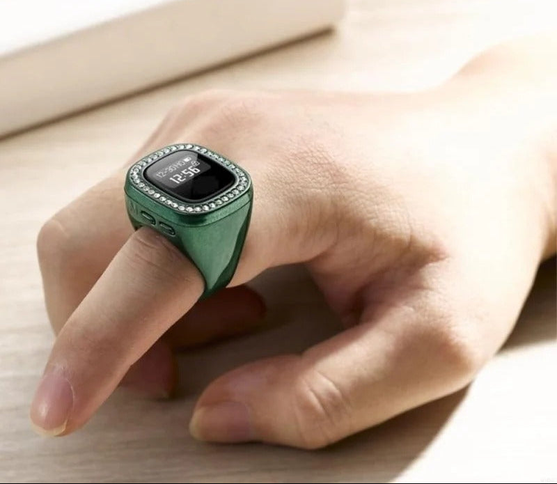 A green smart ring with a rhinestone-studded bezel worn on a person's finger, shown against a light wood surface with a blurred book in the background. The BIN ZAHID MAQET logo is visible in the top-left corner.